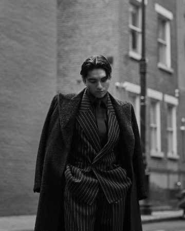 Man wearing a pinstripe suit and overcoat outdoors on a city street in a black and white fashion editorial photograph.