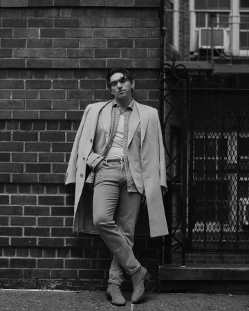Man wearing a tailored coat and denim jeans leaning against a brick wall in a black and white editorial portrait.