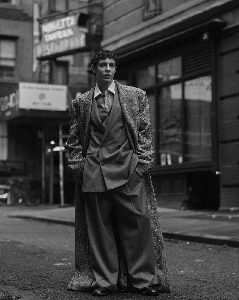 Monochrome portrait of Ben Levi Ross wearing an oversized tweed coat and tailored suit on a city street.