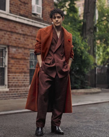 A young man standing outdoors on a street wearing a rust orange overcoat and a patterned burgundy double-breasted suit.