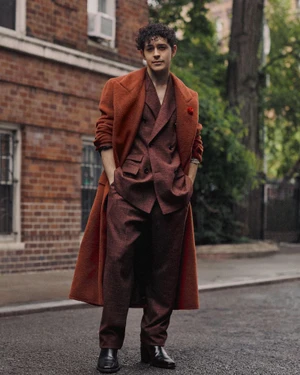 A young man standing outdoors on a street wearing a rust orange overcoat and a patterned burgundy double-breasted suit.