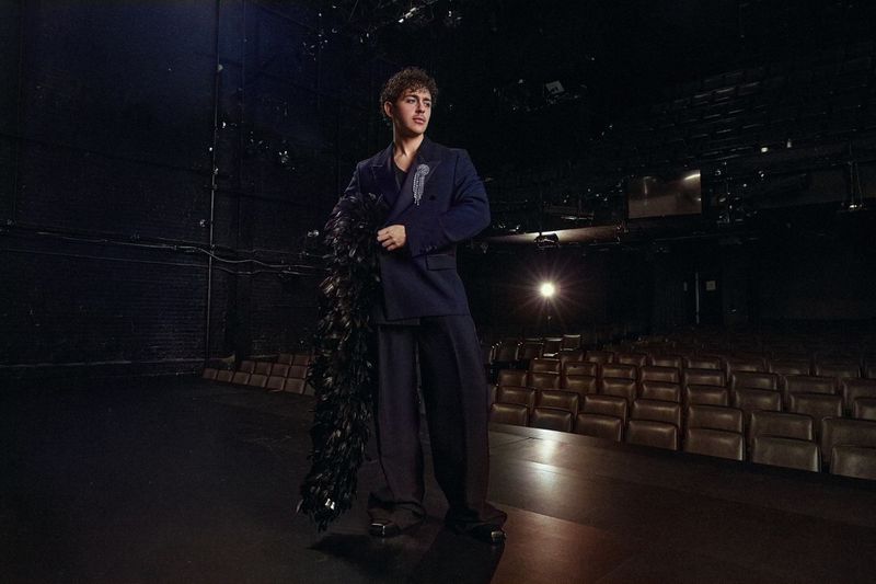 Man wearing a dark suit holding a black feather boa standing on a dark theatre stage for V Man