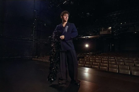 Man wearing a dark suit holding a black feather boa standing on a dark theatre stage for V Man