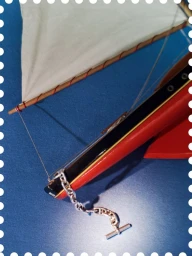 Model sailboat bow with red and black hull, white sail, and silver chain detail on a blue surface