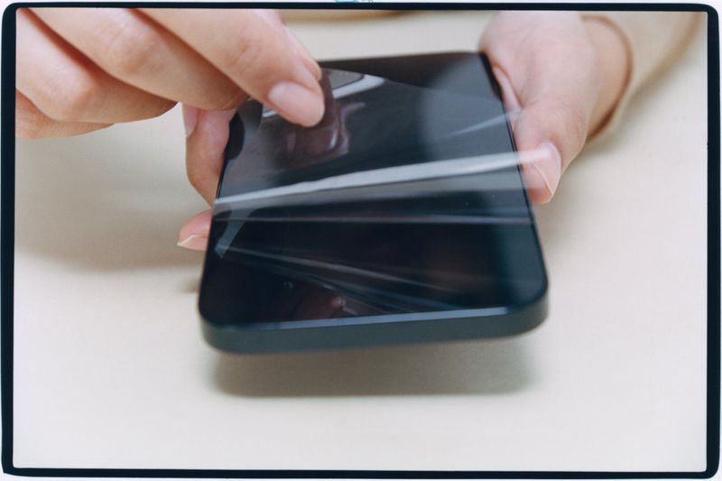 Close-up of hands peeling a protective plastic film off the screen of a dark smartphone.