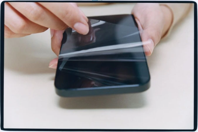 Close-up of hands peeling a protective plastic film off the screen of a dark smartphone.