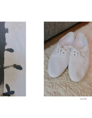 A pair of white leather lace-up shoes resting on a textured white rug next to an abstract black and white art panel.