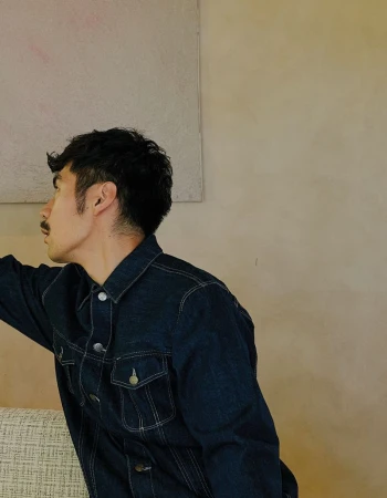 Man with dark hair and mustache wearing a dark denim jacket in profile view against a neutral wall