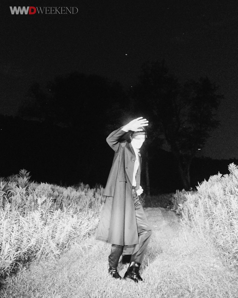 Monochrome night photograph of a man wearing a dark trench coat walking along a grassy path with trees.