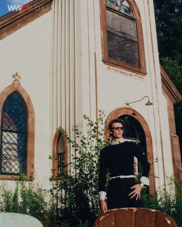 Model wearing dark sunglasses and a black sweater standing before a light-colored architectural building exterior.