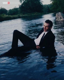 Man wearing a black suit and white shirt reclining in shallow flowing river water for WWD editorial
