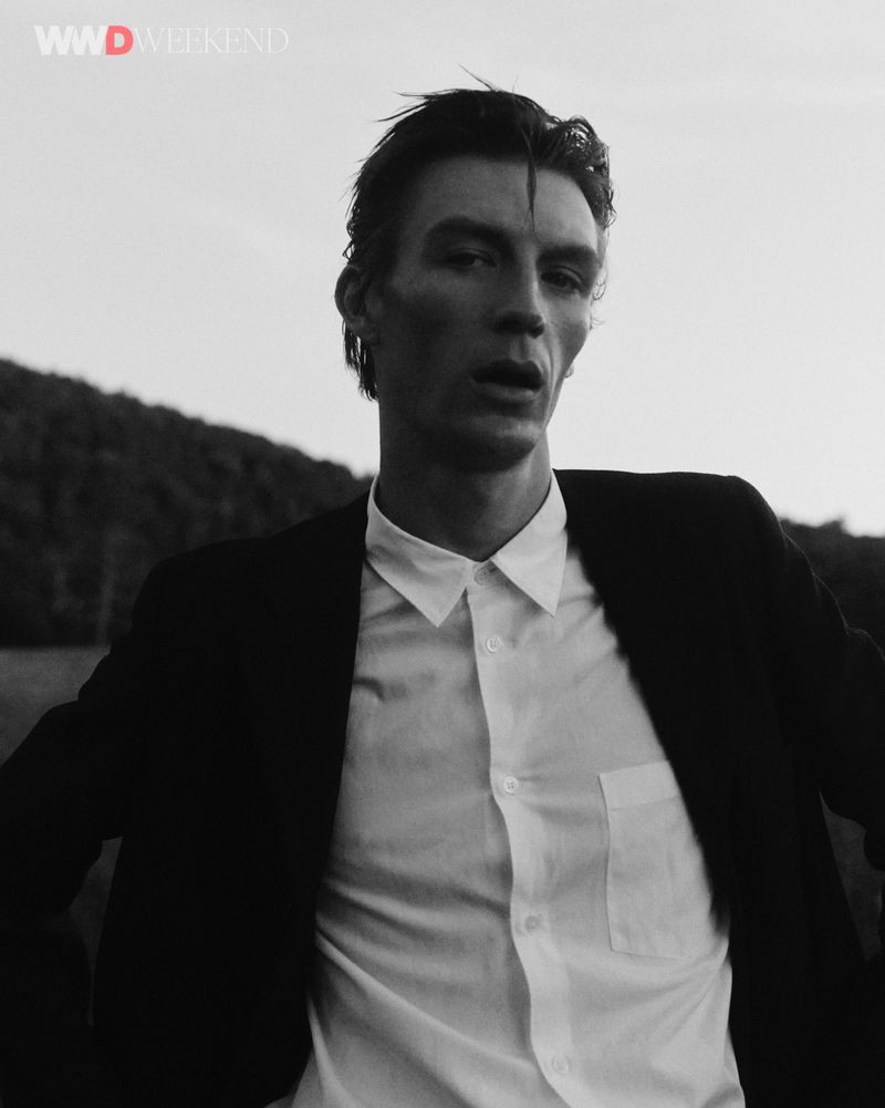 Man wearing a dark blazer over a white button-down shirt outdoors against a natural landscape background.