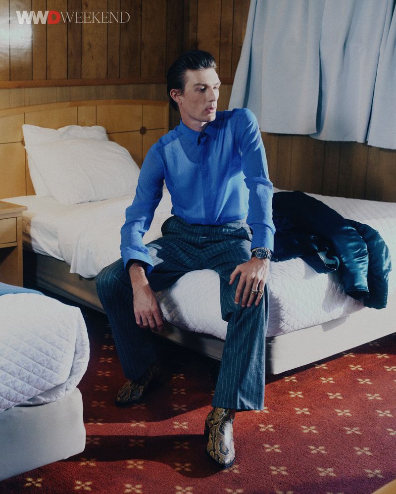 Male model Finnlay Davis wearing a blue shirt and grey pinstripe trousers seated on a hotel room bed for WWD.