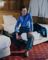 Male model Finnlay Davis wearing a blue shirt and grey pinstripe trousers seated on a hotel room bed for WWD.