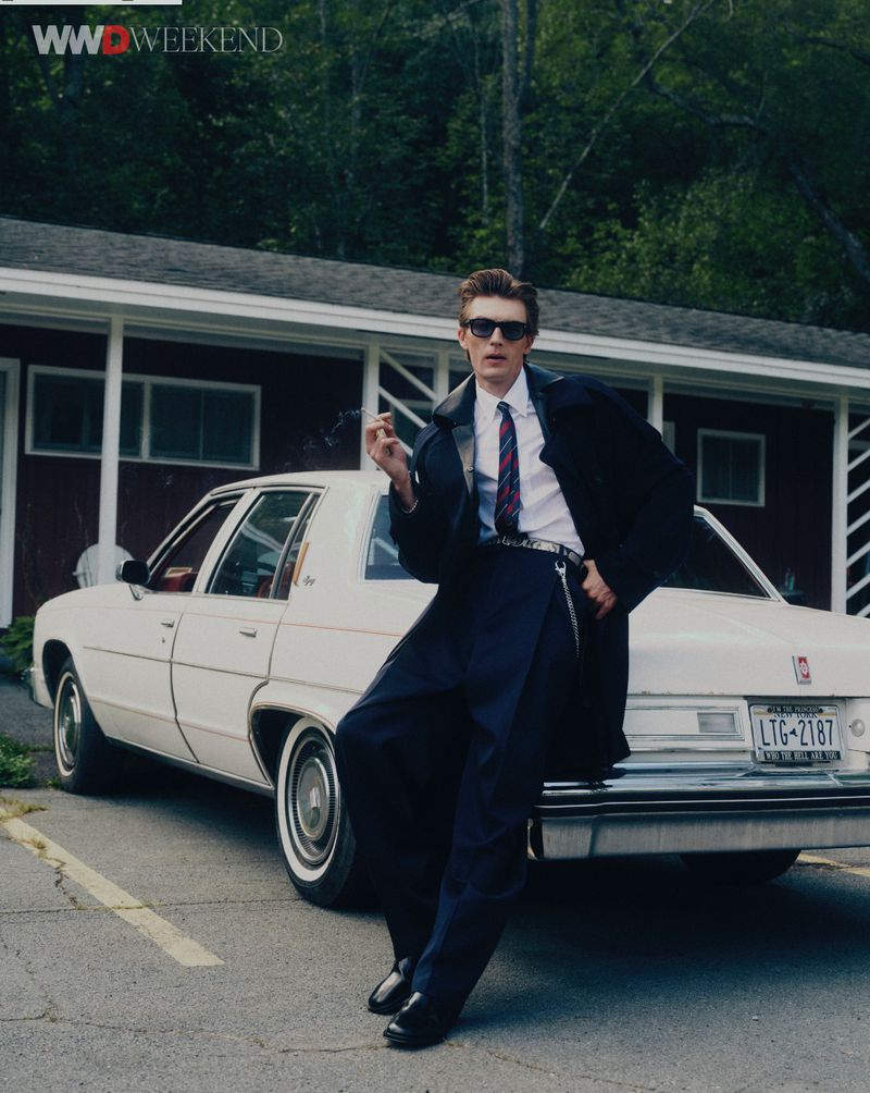 Model leaning against a vintage white car in front of a motel building, wearing a dark coat and sunglasses.