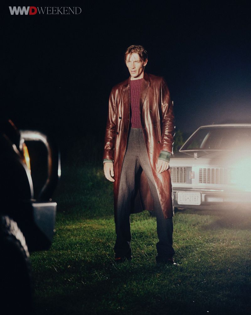 Man in a brown leather coat standing outdoors at night with a car's headlights on in the background.