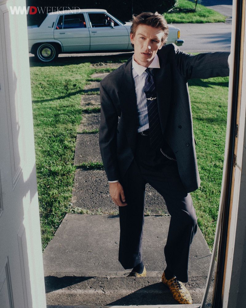 Model wearing dark suit standing on concrete steps with a vintage car parked in the background