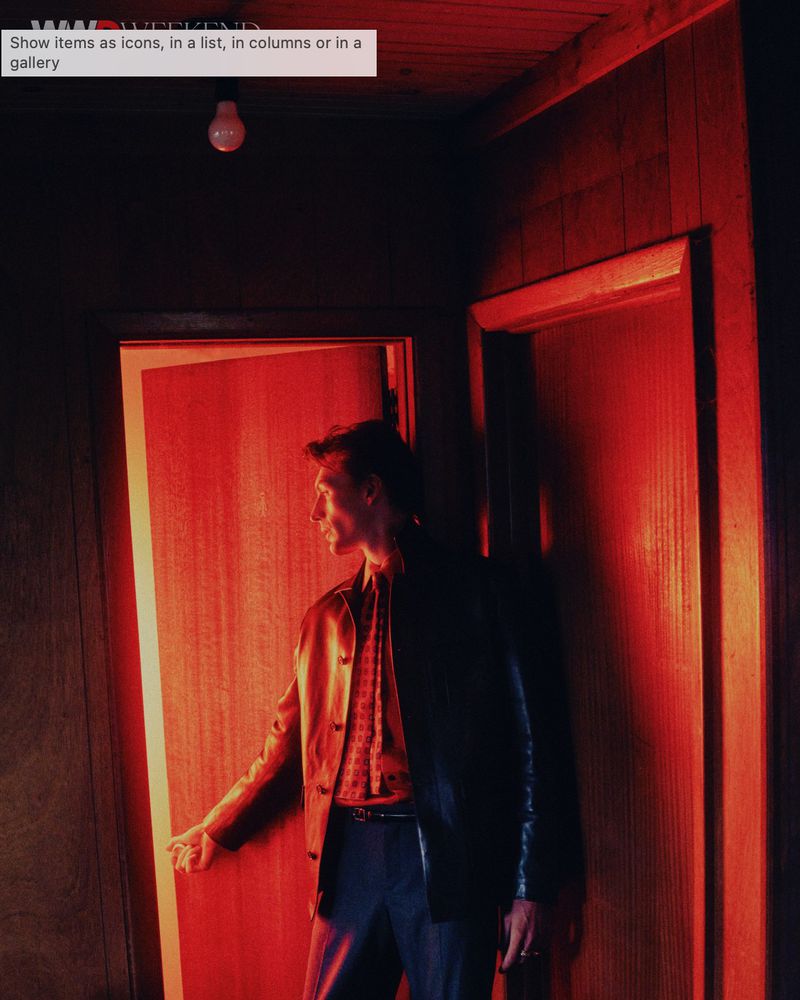 A man in a dark jacket and patterned shirt standing in a red-lit room opening a doorway for the WWD Neo Noir editorial project.