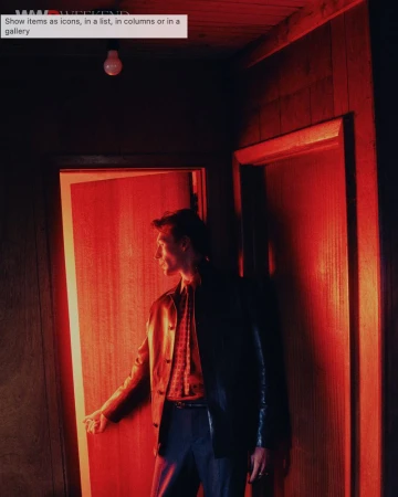 A man in a dark jacket and patterned shirt standing in a red-lit room opening a doorway for the WWD Neo Noir editorial project.