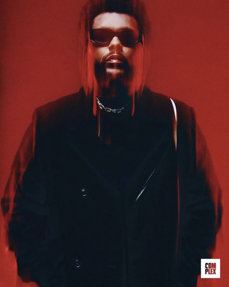 Adult male with a beard and sunglasses, wearing a dark coat and chain necklace, poses against a vibrant red studio backdrop.