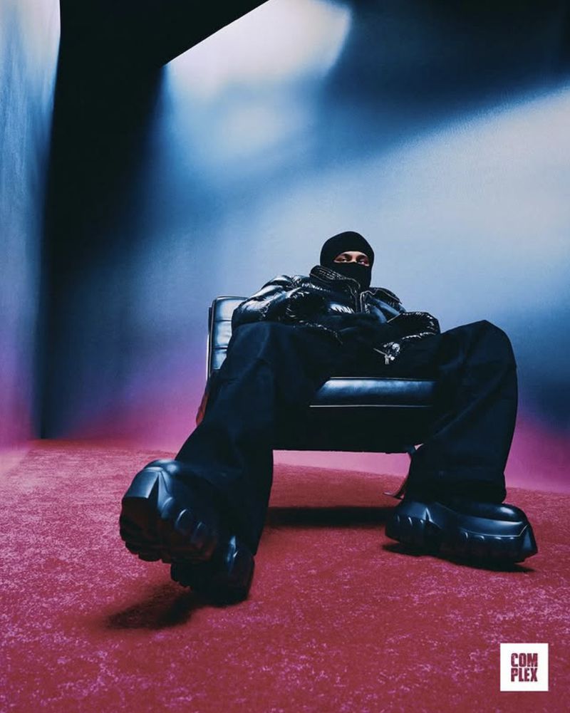 Adult in a balaclava and jacket sits in a chair in a photo studio with dramatic lighting and a textured carpet.