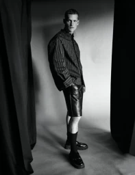 Model in striped shirt and leather shorts standing in a studio framed by dark curtains for Esquire Portugal