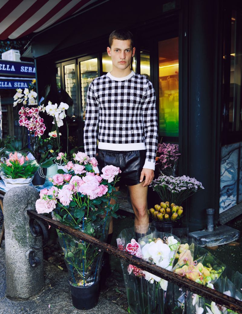 Model wearing a black and white gingham sweater standing amongst flower arrangements outside a storefront.