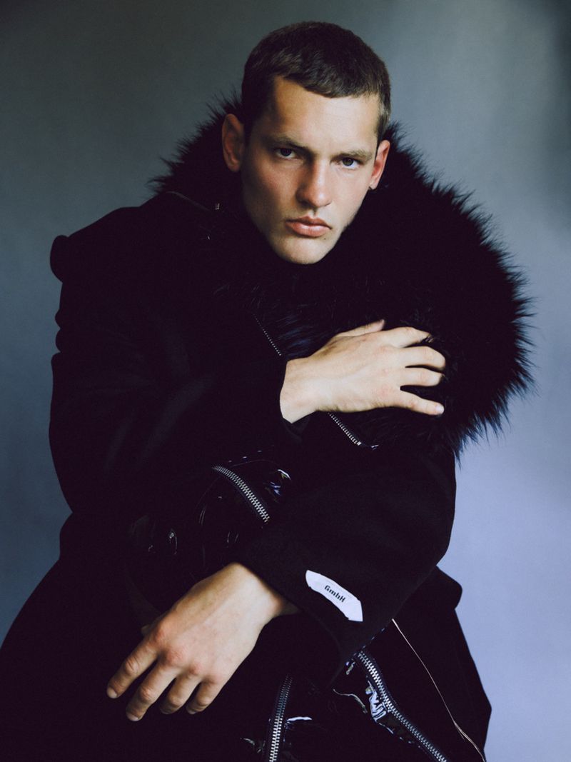 Close-up portrait of a male model wearing a heavy black faux fur collared jacket against a plain grey studio backdrop.