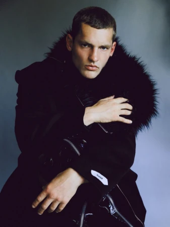 Close-up portrait of a male model wearing a heavy black faux fur collared jacket against a plain grey studio backdrop.