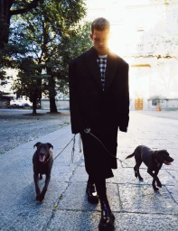 Man wearing a black coat walking two dogs on leashes on a stone paved square with bright sunlight