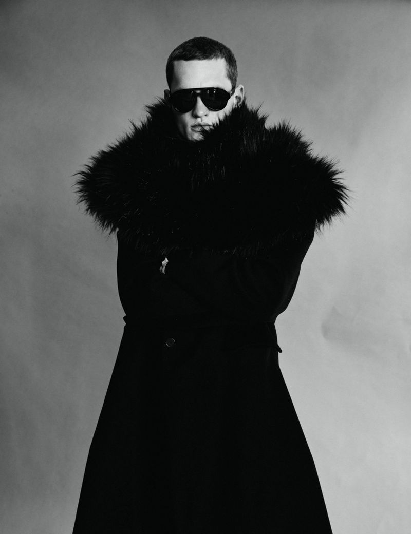 Monochrome portrait of a male model wearing dark sunglasses and a heavy faux fur collar coat against a plain background