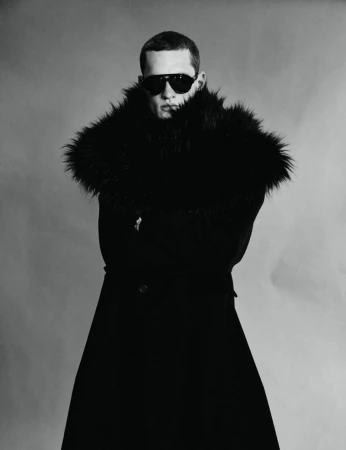 Monochrome portrait of a male model wearing dark sunglasses and a heavy faux fur collar coat against a plain background