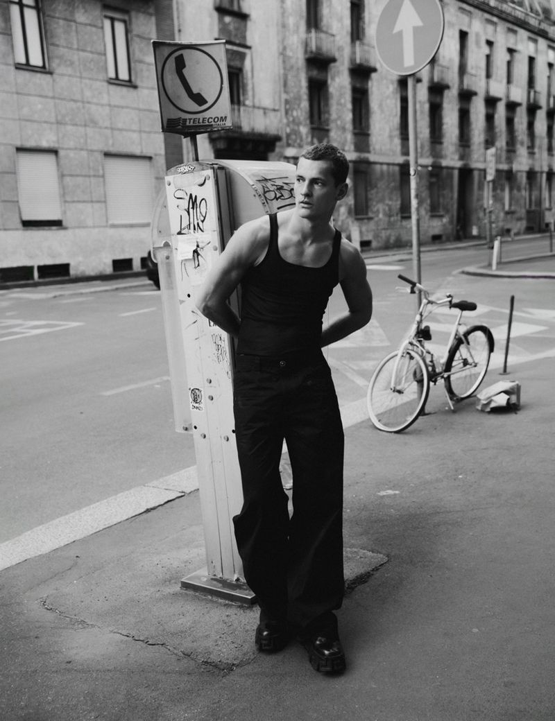 Man wearing a black tank top and trousers leaning against a street sign post on a city street.
