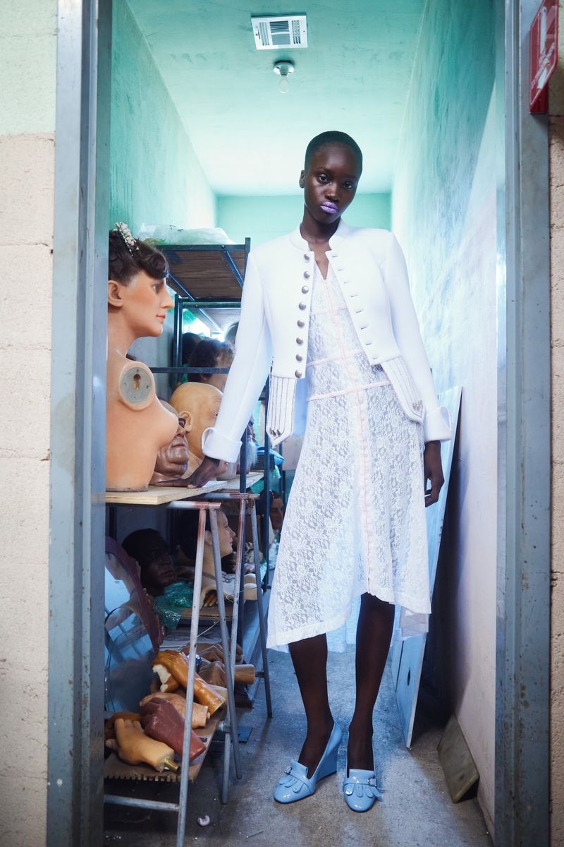 Black model in a white lace dress and white jacket standing in a narrow workshop filled with mannequin heads