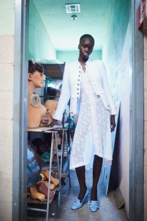 Black model in a white lace dress and white jacket standing in a narrow workshop filled with mannequin heads