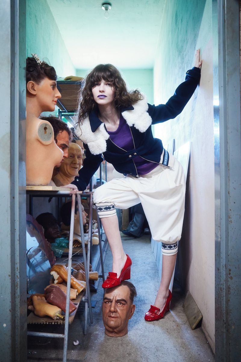 Model in dark jacket with white fur collar and white trousers placing red wedge shoe on a mannequin head