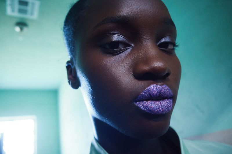 Close-up portrait of a Black woman with shimmering purple glitter lipstick and glossy eyeshadow against a teal wall