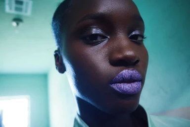Close-up portrait of a Black woman with shimmering purple glitter lipstick and glossy eyeshadow against a teal wall