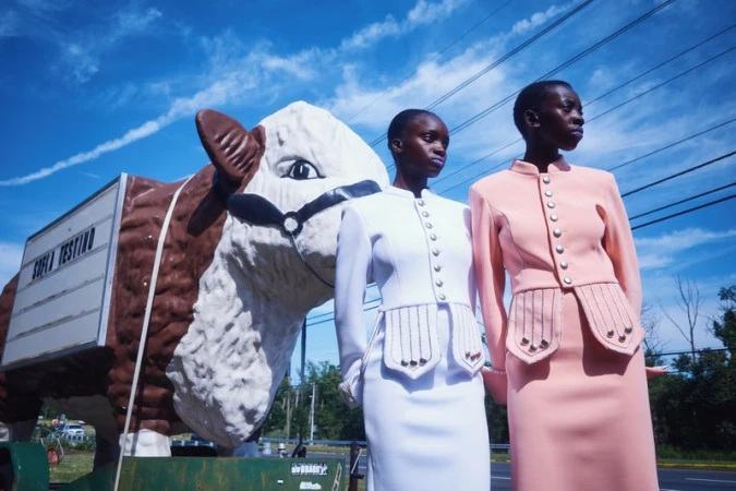 Two black women in matching buttoned jackets and skirts standing outdoors beside a giant white and brown bull statue.