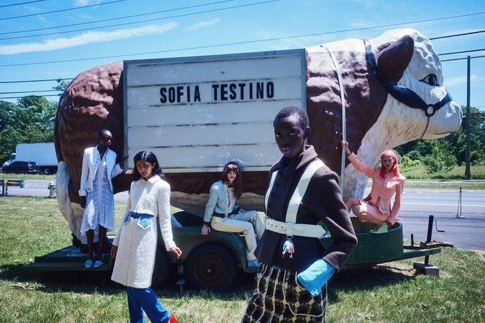 Five models posing near a giant roadside cow statue with a sign reading Sofia Testino for an AnOther Magazine editorial.