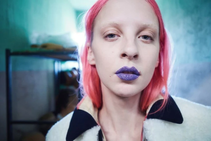Close-up portrait of a woman with vibrant pink hair and sparkling purple lipstick wearing a textured white jacket with a dark collar.