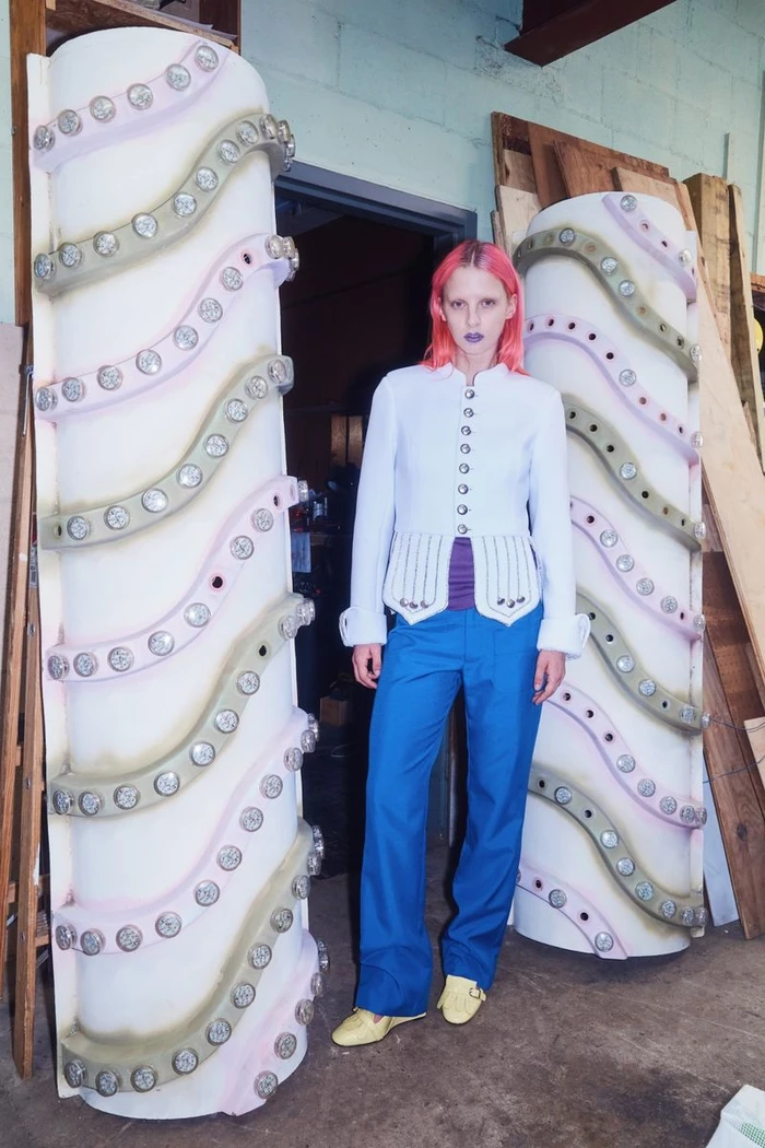 Model with pink hair standing between two large cylindrical columns wearing a white button jacket and blue trousers