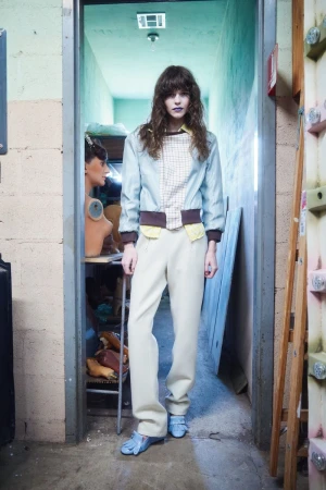 Model standing in a doorway wearing a light blue jacket, patterned top, off-white trousers, and blue loafers.