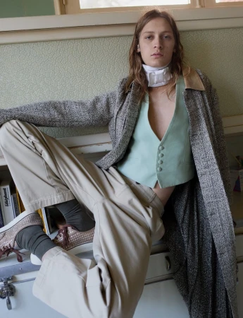 Model wearing a tweed coat over a mint green vest and beige trousers, posed indoors for Replica Man Magazine.