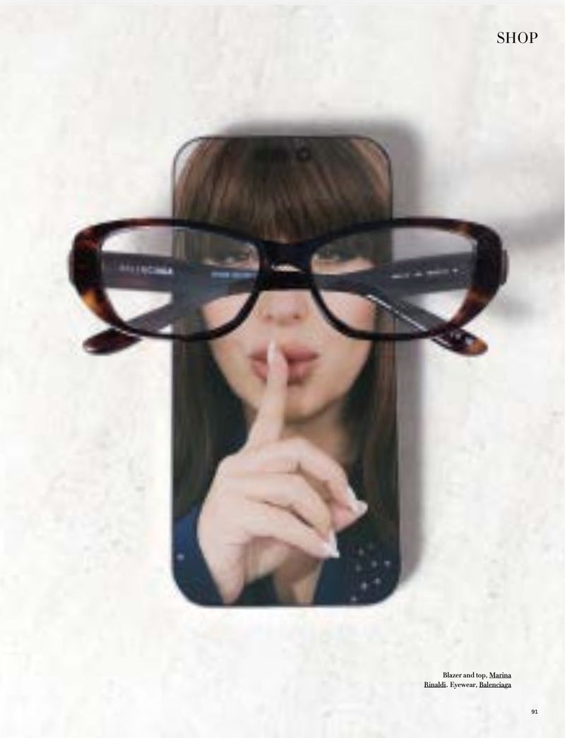 Close-up portrait of a woman holding a smartphone displaying her face, wearing tortoiseshell glasses.