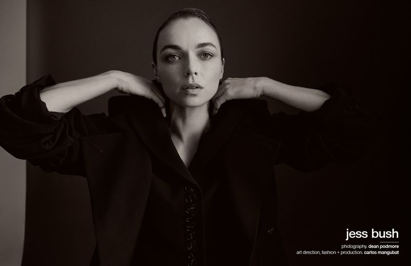 Portrait of actress Jess Bush wearing a dark jacket with her hands near her shoulders against a neutral dark studio background.