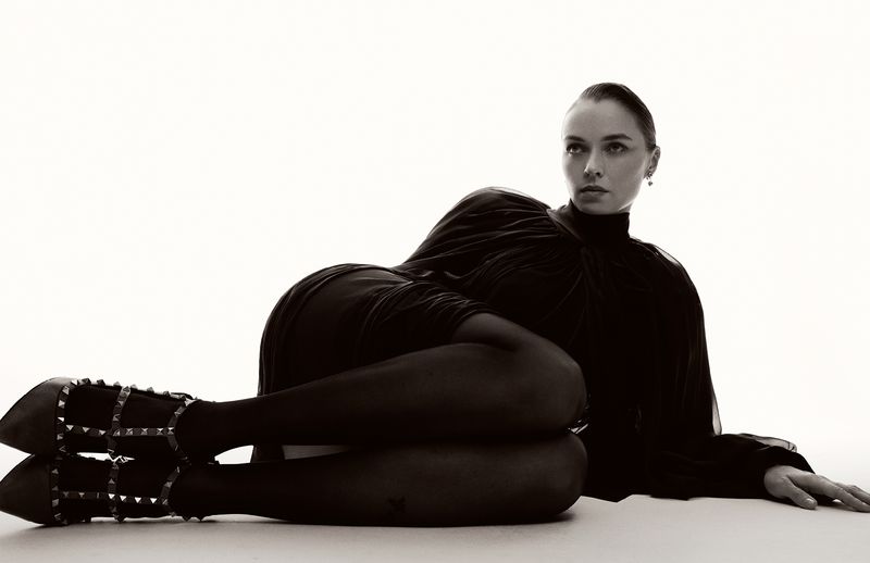 Model Jess Bush lying down in a dark dress and heeled shoes against a bright white background for Schön Magazine interview feature.