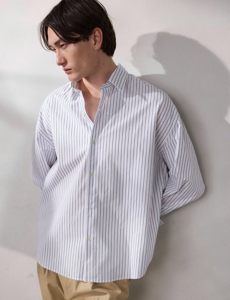 Model wearing a white striped button-down shirt and beige trousers leaning against a white studio wall.