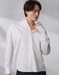 East Asian man in a loose white button-down shirt adjusting his collar against a textured light grey backdrop.