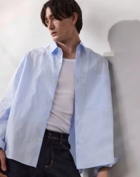 Model wearing an unbuttoned light blue shirt over a white tank top and dark jeans against a neutral wall.